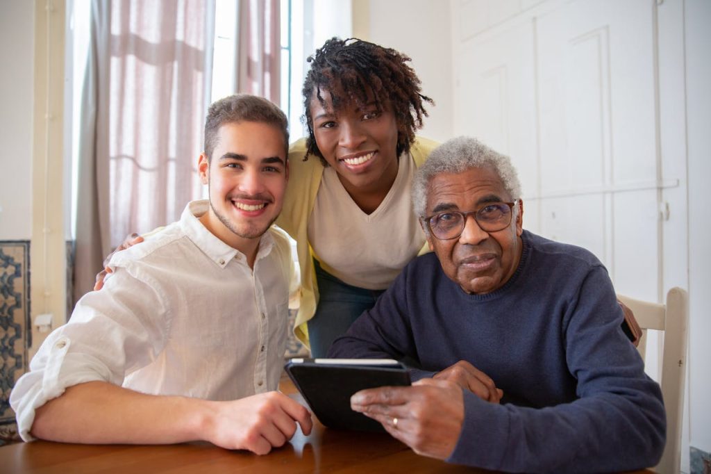 a-group-of-people-smiling-together-while-sitting-near-the-table-7551617 Elderly man enjoying technology with diverse caregivers at home.
