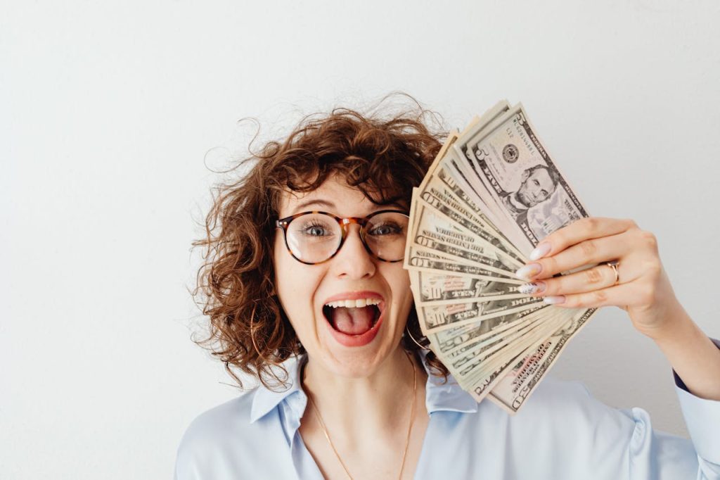 happy-woman-in-blue-long-sleeve-blouse-holding-money-7680637 Caucasian woman with curly hair delights in holding US dollars against a white background.